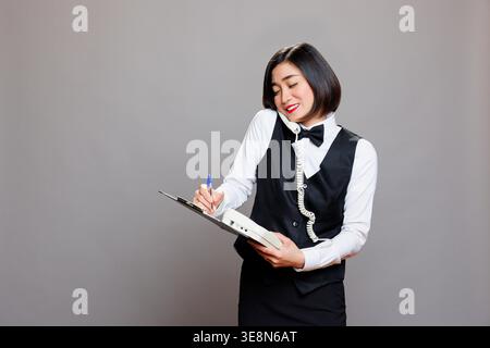 Smiling asian waitress answering landline phone call and writing on clipboard. Restaurant friendly woman employee in uniform talking with client on telephone and managing order Stock Photo
