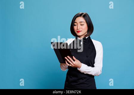 Restaurant young waitress using digital tablet while managing customer order online. Attractive hotel woman receptionist in uniform tapping on portable device touchscreen Stock Photo