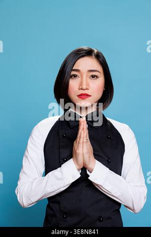 Young asian woman receptionist meditating with folded palms, relaxing and looking at camera. Attractive cafe waitress wearing uniform standing in zen pose portrait on blue background Stock Photo