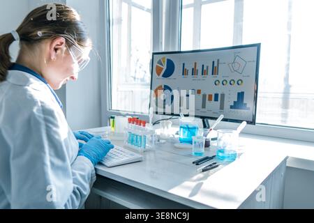 Young researcher wearing protective eyewear, gloves works on keyboard in sunlit laboratory. Data analysis, scientific research, laboratory experiments are highlighted in clean, high tech environment Stock Photo