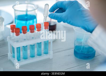 A close up view of laboratory research shows a gloved hand handling a pipette with blue liquid. Test tubes, beakers, and scientific equipment are visible in a clean, bright lab setting Stock Photo