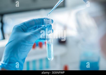 Close up of a gloved hand holding a test tube while using a pipette to add blue liquid in a laboratory setting. Focus on scientific experiment, biotechnology, and innovation Stock Photo