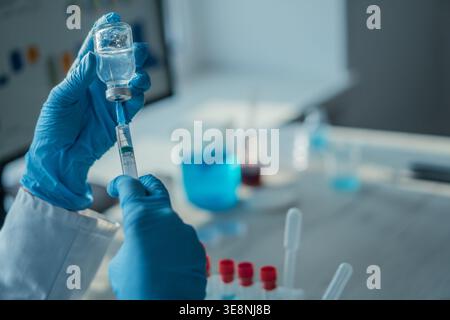 Close up of hands in blue gloves preparing an injection with a syringe and vial, surrounded by laboratory equipment. The image emphasizes medical research, vaccination, and scientific innovation Stock Photo