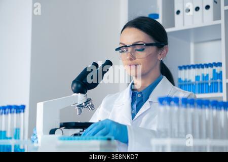 In a wellequipped lab, a dedicated female scientist in a white coat examines samples under a microscope, surrounded by colorful test tubes, showcasing her commitment to biology and chemistry Stock Photo