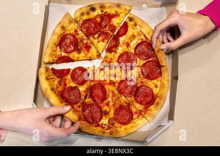 Pepperoni pizza slices shared from takeaway box. Top view of hands reaching for pepperoni pizza slices in a cardboard box, showing casual sharing Stock Photo