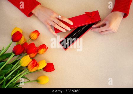 Opening jewelry gift box with tulips. Top view of hands opening a red jewelry box with a necklace beside colorful tulips, suggesting romance Stock Photo