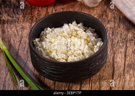 A black bowl of fresh, grainy cottage cheese on a rustic wooden table. Close up view with soft lighting highlighting textures. Green onion tomato in t Stock Photo