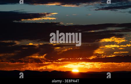 As the sun descends behind the horizon, vibrant hues of orange, red, and purple fill the sky. Soft clouds catch the fading light, casting shadows over Stock Photo