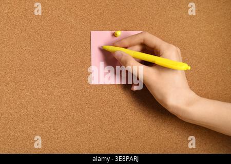 Young woman writing on sticky note on cork board as background Stock Photo