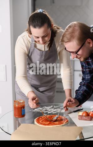 Close up young couple focused on spreading tomato sauce on pizza dough at glass table in kitchen. Man and woman in apron cooking together at home. Culinary process and teamwork concept, domestic hobby Stock Photo