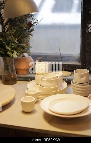 Beautiful still life of a complete handmade ceramic dishware set — plates, cups and bowls — on a bright studio windowsill with a plant. Stock Photo