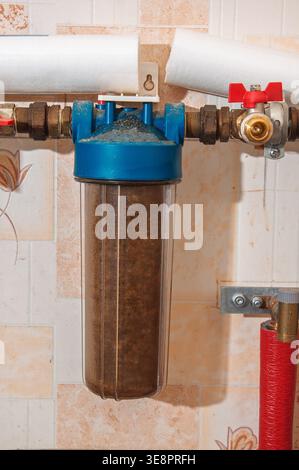 A close-up view of a used, clogged water filter housing filled with sediment and debris, attached to plumbing pipes Stock Photo