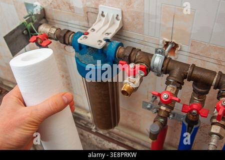 A hand holds a new white filter cartridge next to an installed, used water filter housing with brass plumbing and red valves Stock Photo