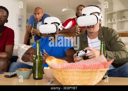Diverse male friends playing VR wearing white headsets holding controllers on couch with beer Stock Photo