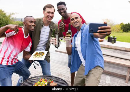 Four men posing and taking selfie on wooden deck with charcoal grill skewers and smartphone Stock Photo