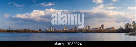 Wide panorama of Obolon embankment in Kyiv, where golden evening light touches the Dnipro River, dramatic clouds drift above the skyline, and the city Stock Photo