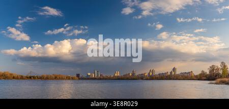 Wide panorama of Obolon embankment in Kyiv, where golden evening light touches the Dnipro River, dramatic clouds drift above the skyline, and the city Stock Photo