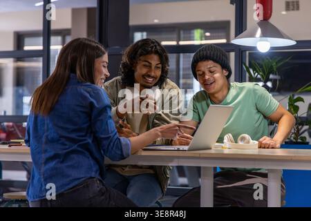 Diverse students leaning in, discussing group project at study lounge with laptop and headphones Stock Photo