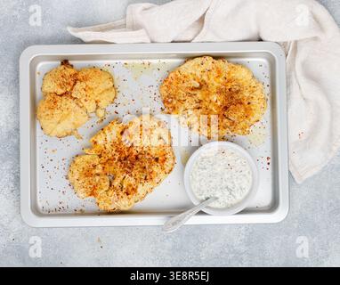 Baked cauliflower steaks with herbs and spices on a baking sheet on a gray concrete background. Mayonnaise sauce with dill, parsley and garlic. Vegan, Stock Photo