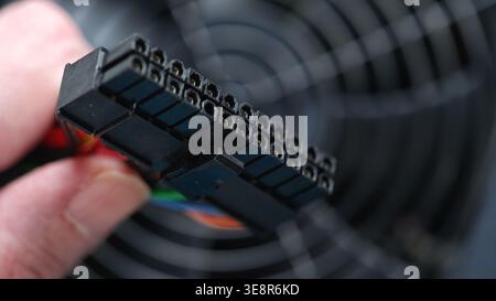 A hand holds a 24-pin ATX power connector with visible dust on its surface against the fan grill of a computer power supply unit. Close up. Stock Photo