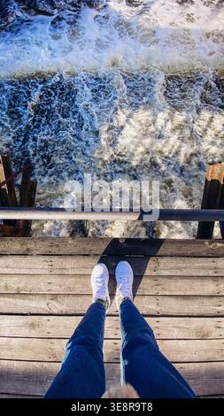 Footsteps are visible on wooden surface above rushing water. Waves crash below creating foam. Sunlight reflects off the water. The setting is busy and Stock Photo