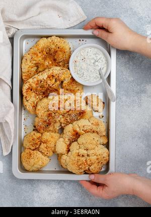 In women's hands, a baking tray with Baked cauliflower steaks with herbs and spices and mayonnaise sauce with dill, parsley and garlic. Vegan, vegetar Stock Photo