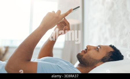 Man Lying on Bed Using Smartphone While Looking at Screen in Bright Room Setting Stock Photo