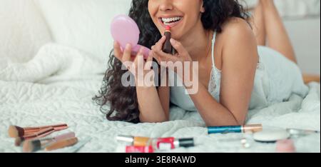 Woman Applying Lipstick While Lying on Bed Surrounded by Makeup Products and Tools Stock Photo