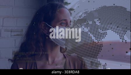 Standing mid adult woman wearing brown sweater, surgical mask by window, showing globe map overlays Stock Photo