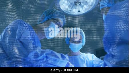 Operating three surgical staff wearing gowns leaning over sterile field in OR, with overhead light Stock Photo