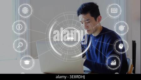 Typing Asian man using silver laptop at desk by window, wearing dark blue sweater, showing HUD Stock Photo