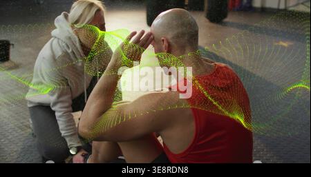 Bald trainee doing crunches on gym floor trainer kneeling bracing feet with tracker, green waveform Stock Photo