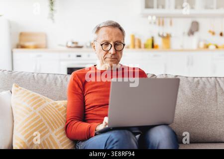 Older Man Sitting on a Sofa Using a Laptop in a Bright Kitchen During Daytime Hours Stock Photo