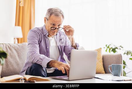 Man Using a Laptop in a Living Room While Adjusting His Glasses and Focusing on the Screen Stock Photo
