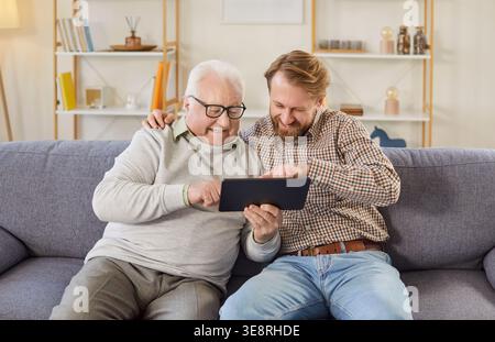 Happy senior father and adult son sitting on sofa, using tablet and enjoying digital leisure time. Stock Photo
