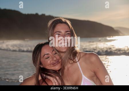 Two cheerful female friends hugging on a beach with ocean waves at golden hour Stock Photo