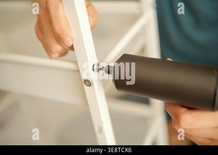 Woman hand using power drill to assemble white metal frame of compact storage carts for kitchen or bathroom at home. Stock Photo