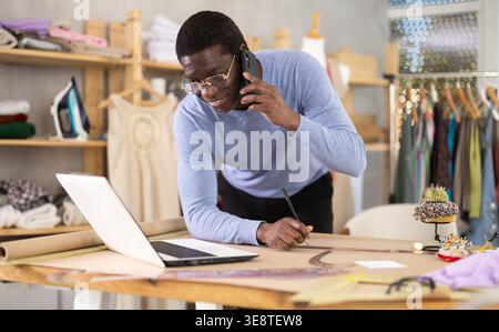 Tailor communicating on phone and reviewing specifications on laptop in workshop Stock Photo