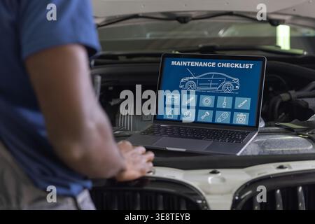 Auto mechanic checks car maintenance checklist on laptop while working on engine in garage. He focuses on vehicle repair tasks. Stock Photo