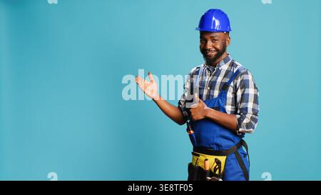 African american repairman pointing at something in studio, indicating let or right direction for construction advertisement. Cheerful male carpenter presenting sideways for studio ad. Stock Photo