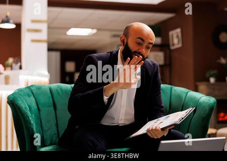 Businessman attends videocall in hotel lobby, killing time while he waits to check in. Entrepreneur in suit using laptop to meet online with manager, videoconference telework in lounge area. Stock Photo