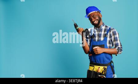 African american man using power drilling tool on wall, drill nails with cordless nail gun on camera. Professional handyman with overalls holding drill gun for holes, construction equipment. Stock Photo