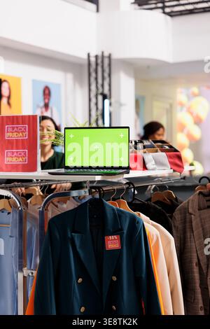 Vertical photo of open laptop with chroma key green screen standing on clothing rack during Black Friday sales in retail store. Inventory management softwares for apparel retail Stock Photo