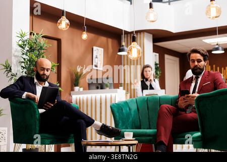 Two businessman in lounge area waiting to do check in process and relax after long flight, travelling on work purposes. White collar workers in hotel lobby preparing for upcoming corporate event. Stock Photo