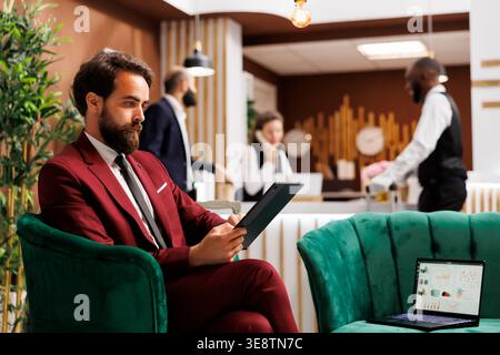 Employee looks over meeting notes waiting to do check in before attending important international conference. Young businessman at hotel travelling for work, reading documents in lobby. Stock Photo