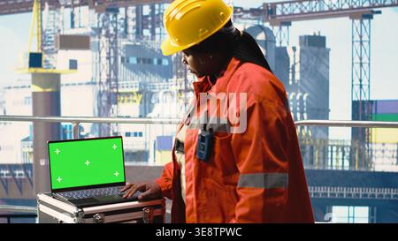 Green screen laptop used by drilling rig worker holding handheld radio device to monitor operation. Woman on offshore platform deck using walkie talkie and mockup notebook, calibrating gear Stock Photo