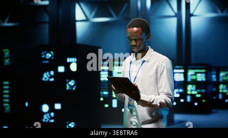 IT programmer in server hub using tablet to find firewall misconfigurations affecting mainframes performance and connectivity, leading to slow data transfer rates and poor client communication Stock Photo