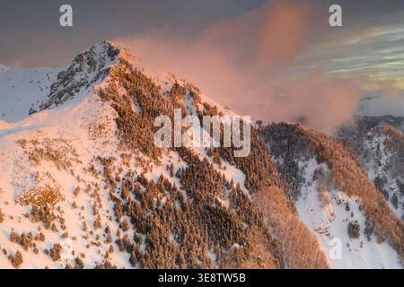 Aerial view of a snow-covered mountain peak with pine trees on the slopes under a pink sunset sky with clouds Sassalbo, Tuscany, Italy. Stock Photo