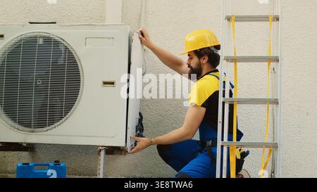 Experienced wireman dissasembling external air conditioner metal side coil panel to check for bad wiring. Meticulous electrician doing repairing on malfunctioning hvac system Stock Photo