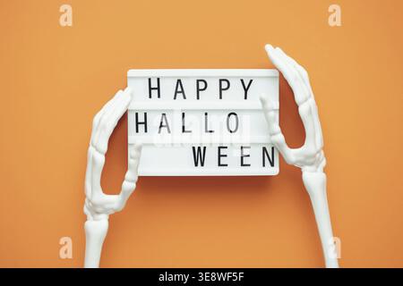 The inscription HAPPY HALLOWEEN on a lightbox on an orange background in the hands of a skeleton. Greeting card for the autumn holiday. The concept of Stock Photo